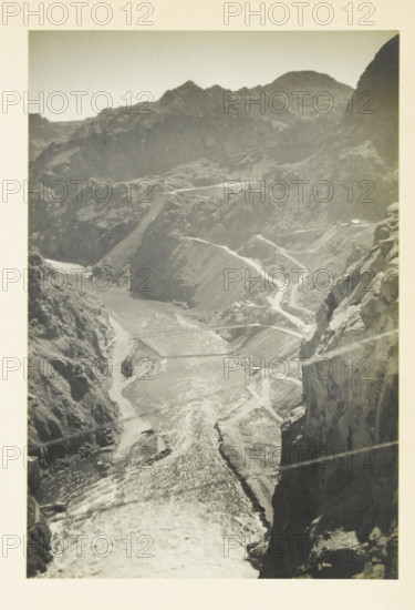 View in the Black Canyon, United States, Black Canyon, photograph, title: 'BLACK CANYON', photographer: attributed to Wouter Cool, United States of America, 1936, North America, Attributed to, pasteboard, paper, gelatin silver print, developing gelatin silver print, height 227 mm x width 153 mm, height 315 mm x width 212 mm, ravine, chasm, abyss, canyon, A tall vertical black-and-white photograph of a deep rocky canyon with steep cliffs on both sides and a winding river flowing through the center, bright reflective water visible along its length, narrow switchback roads carved into the canyon walls, layered rugged mountain ridges in the background under a pale sky, and rough textured rock faces with visible striations and ledges in the foreground.