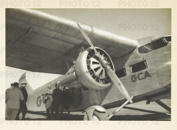 Our plane for the flight over the Grand Canyon, Aircraft before departure for a flight over the Grand Canyon, United States, photograph, title: 'OUR PLANE FOR THE, FLIGHT OVER THE GRAND CANYON', photographer: attributed to Wouter Cool, United States of America, 1936, North America, Attributed to, pasteboard, paper, gelatin silver print, height 154 mm x width 226 mm, height 315 mm x width 285 mm, aircraft, aeroplane, A black and white photograph showing a close-up of a corrugated metal airplane wing and fuselage with a radial engine and propeller mounted beneath the wing, the engine cowling and strut visible with ribbed metal texture, a portion of the cockpit window and passenger windows on the fuselage, the landing gear strut and wheel at the bottom, and a group of people standing beside the plane near the entrance, some wearing hats and coats.