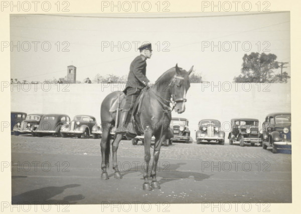 Police officer on horseback, Hoboken pier, New York, United States, Traffic officer in front of the pier, photograph, title: 'TRAFFIC OFFICER, IN FRONT OF THE PIER.', photographer: attributed to Wouter Cool, United States of America, 1936, North America, Attributed to, pasteboard, paper, gelatin silver print, height 155 mm x width 227 mm, height 315 mm x width 286 mm, policeman, constable, pier, quay, wharf, A black and white photograph showing a person in a uniform and cap seated on a dark horse in the foreground, the horse facing slightly toward the camera with visible tack and a long tail, and a row of vintage cars parked in front of a high light-colored wall behind them, with part of a small tower and some trees visible above the wall and a paved surface under the horse and cars.