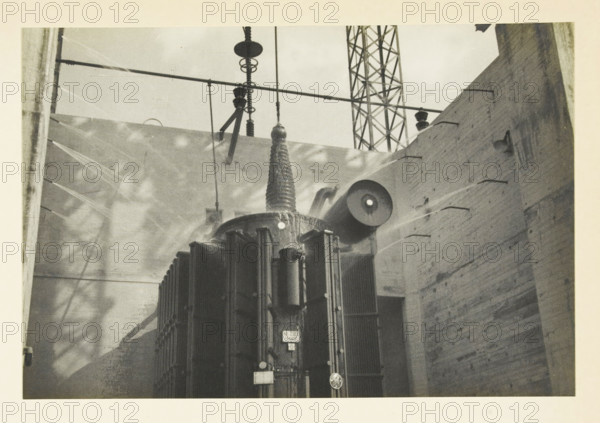 Safe Harbor Dam, Pennsylvania, United States: fire extinguishing system, Fire extinguishing system of transformers at Safe Harbor, photograph, title: 'FIRE EXTINGUISHING INSTALLATION OF, TRANSFORMERS AT SAFE HARBOR.', photographer: attributed to Wouter Cool, United States of America, 1936, North America, Attributed to, pasteboard, paper, gelatin silver print, developing gelatin silver print, height 153 mm x width 227 mm, height 315 mm x width 330 mm, dam (in canal), A large industrial transformer with vertical cooling fins and cylindrical attachments sits between two tall brick walls, with insulators and suspension hardware extending upward from its top toward horizontal crossarms; a lattice steel tower is visible in the background, and light and shadow patterns fall across the walls and transformer surfaces.