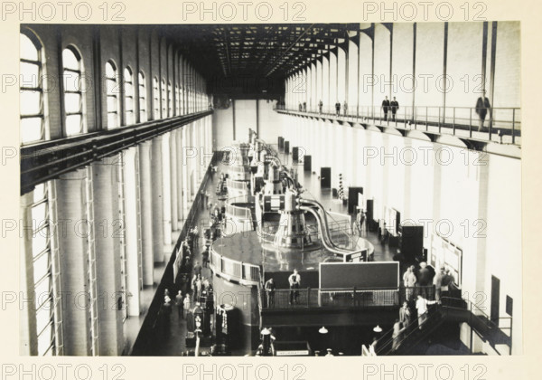 Safe Harbor Dam, Pennsylvania, United States: interior of the power station, Interior Safe Harbor power station, photograph, title: 'INTERIOR POWER PLANT, SAFE HARBOR.', photographer: attributed to Wouter Cool, United States of America, 1936, North America, Attributed to, pasteboard, paper, gelatin silver print, developing gelatin silver print, height 153 mm x width 231 mm, height 315 mm x width 285 mm, dam (in canal), A large interior space with high arched windows along the left wall and tall vertical supports along the right, a long overhead catwalk with railings and several people walking on it, a row of large cylindrical machines with curved metal piping and railings down the center floor, groups of people standing and walking around the machines and along the floor, staircases and railings leading between levels, exposed ceiling trusses and beams above, and display boards and panels positioned near the right wall.
