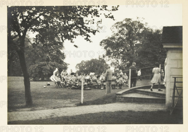 Military Chapel at Mount Vernon, Mount Vernon, Washington, United States: military chapel, photograph, title: 'MILITARY CHAPEL AT, MOUNT VERNON.', photographer: attributed to Wouter Cool, United States of America, 1936, North America, Attributed to, pasteboard, paper, gelatin silver print, developing gelatin silver print, height 153 mm x width 225 mm, height 315 mm x width 285 mm, parts of church exterior and annexes: chapel - QQ - small church, chapel, A black and white photograph of an outdoor scene showing a seated brass band with musicians holding instruments, music stands, and a large brass tuba, arranged on folding chairs on a grassy area beneath trees; several people stand nearby watching, including a man in a suit and a girl in a dress walking up steps toward a small building with a tiled roof and a visible corner wall; a low rope or barrier runs between posts in front of the band, and paved walkways and a short brick ledge are visible in the foreground, with trees and open sky...