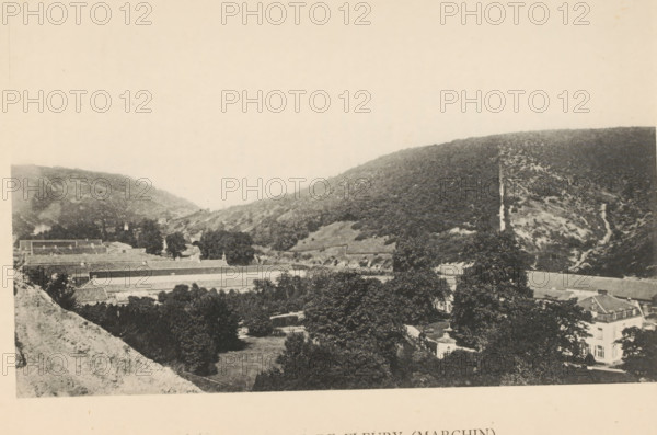Les Papeteries de Fleury (Marchin), View of the paper mills of Marchin, page, photomechanical print, photographer: attributed to Victor Gaillard, Marchin, cliché maker: attributed to Emile Aubry, Brussels, c. 1885 - in or before 1890, Attributed to, paper, collotype, photoprint, height 104 mm x width 159 mm, factory in landscape, A sepia-toned landscape showing a valley flanked by wooded hills, with terraced rooftops and long low buildings visible along the hillside and in the valley floor; scattered trees and a manicured lawn occupy the foreground, and a larger multi-story building with a pitched roof appears at the right edge, while paths and a narrow lighter strip run up the slope on the far hill.