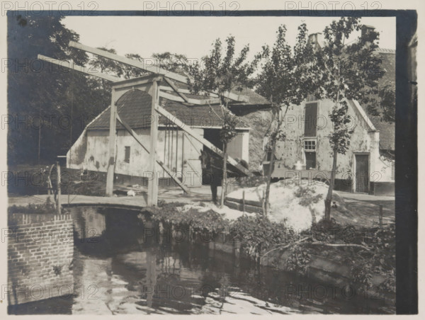 Bij Voorburg, photograph, Farm with horse and drawbridge, photographer: HC Wilkens (signed by artist), Voorburg, 1915, paper, gelatin silver print, developing gelatin silver print, height 90 mm x width 122 mm, bridge, A riverside scene showing a small bridge and a wooden drawbridge structure with long beams and supporting posts in front of buildings with tiled and thatched roofs, rough plastered walls, and varied door and window openings; a person stands near the bridge beside a horse partially obscured by the structure; young trees with leaves grow along the bank and in front of the buildings; a mound of light-colored material sits near the water edge beside a wheel and other scattered objects; the water in the foreground reflects the bridge, beams, and nearby foliage, and a brick wall lines part of the riverbank.