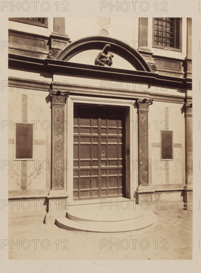 Portal with relief of Mary and Child in the round arch above, photograph, c. 1880 - c. 1900, photographer, pasteboard, paper, albumen print, albumendruk, height 358 mm x width 272 mm, height 650 mm x width 500 mm, closed door, A stone facade with decorative pilasters and carved capitals flanks a large recessed wooden door composed of square panels, set above curved stone steps; above the doorway an arched pediment contains a sculpted figure of a seated person holding a smaller figure, and rectangular framed panels are visible on the surrounding wall surfaces, with ornamental mouldings and a gridded window in the upper corner.