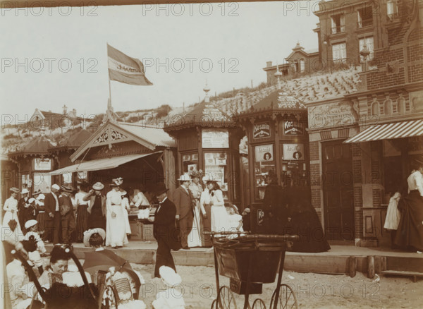 At the Beach, photograph, photographer: attributed to Willem de Jong, Scheveningen, photographer: attributed to Piet de Jong, c. 1895 - c. 1920, Attributed to, paper, baryta paper, daylight collodion silver print, height 89 mm x width 120 mm, open-air recreation, beach, A crowded seaside scene with a row of ornate wooden kiosks and a brick building along a boardwalk in front of grassy dunes and houses; a large flag flies above one of the kiosks, and decorative roof finials and carved trim appear on the structures; many people in period clothing stand and walk in the foreground, including women in long dresses and hats, men in suits and hats, and children sitting on the sand; a man in dark clothing carries a tray with cups, and a pram with large wheels is visible near the front; awnings extend over some shop fronts and display windows show goods inside.