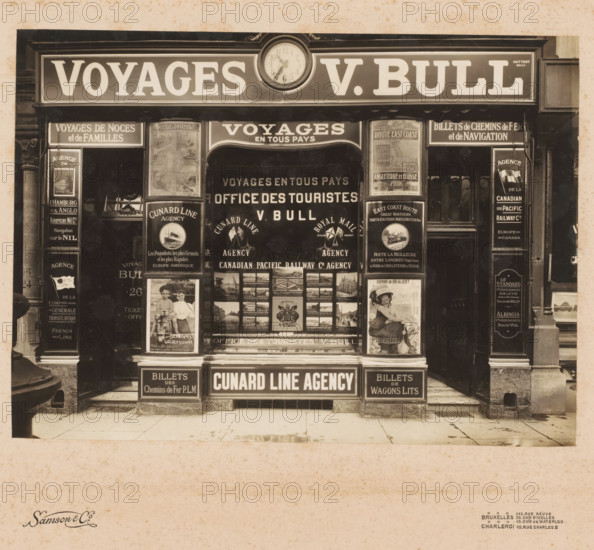 Shop window of the travel agency Voyages v. Bull in Brussels, Belgium, photograph, inscription: 'H. GENEAU, 78, Rue Chevalier, MONTMORENCY (S-&-0)', address: 'BRUXELLES 145, RUE NEUVE, 35, CHÉE D'IXELLES, 13, CHÉE DE WATERLOO, CHARLEROI 10, RUE CHARLES II', photographer: Samson & Cie., Brussels, 1900 - 1910, Samson & Cie., photographer, paper, pasteboard, gelatin silver print, developing gelatin silver print, height 300 mm x width 358 mm, shop-window, show-window, A storefront with a large rectangular sign above and a round clock centered at the top, flanked by display windows filled with framed posters and images, columns on either side of the entrance, a recessed central doorway with glass panes showing more posters inside, small steps leading up from the sidewalk, decorative molding and trim around the facade, and a broad vertical panel area below the windows at street level.