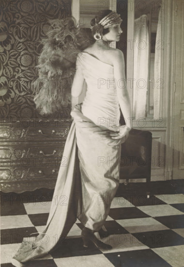 Fashion photo of a woman in a dress by Paul Poiret, Paris, photograph, inscription: '(stamp) P.Pon DE PAUL POIRET, (stamp) PAUL POIRET, 3 MARS 1923, 26, AVENUE ..., 107 FAUBOURG St. ..., (pencil and pen): 3502 Milo ?, ...', after design by Paul Poiret, 1923, photographer, paper, baryta paper, gelatin silver print, developing gelatin silver print, height 172 mm x width 122 mm, Person, model, mannequin, A black-and-white photograph of a woman standing with her back partially turned to the camera, wearing a one-shoulder draped gown that falls into a long train she holds with one hand, a decorative headband across her short hair, heeled shoes visible beneath the gown, a large feathered accessory hanging behind her, patterned wallpaper and a carved wooden chest of drawers to her side, a framed mirror or doorway and a curtain behind her, and a checkerboard floor beneath her feet.