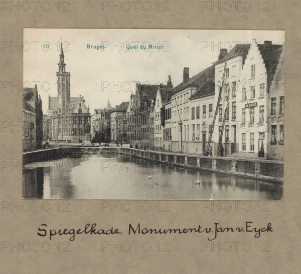 Bruges. Spiegelkade. Monument Jan van Eyck, photograph, 1860 - 1925, photographer, paper, collotype, photoprint, height 100 mm x width 150 mm, height 300 mm x width 250 mm, Person, Jan van Eyck, Bruges, A vintage black and white photograph shows a canal lined by old buildings on one side and a stone embankment on the other, with calm water reflecting parts of the scene and a few swans on the surface; the row of buildings features varied rooflines, stepped gables, many windows, and narrow facades, with ladders leaning against one façade and a small group of people near the canal edge; in the distance a tall ornate tower with a spire rises above a large ornate building with Gothic-style details, and a stone bridge and statues are visible along the canal.