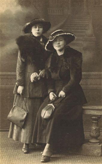 Portrait of two women, photograph, postcard, Germany (possibly), c. 1920 - c. 1930, photographer, paper, gelatin silver print, developed gelatin silver print, height 131 mm x width 81 mm, anonymous historical person portrayed - BB - woman, Two women wearing long dark coats with fur collars and wide-brimmed hats stand and sit in front of a painted backdrop showing steps and foliage; the standing woman holds a small handbag and a floral accessory pinned at her waist while the seated woman rests a hat on her lap and has her hands clasped over it, both wearing ankle boots and facing the camera with neutral expressions.