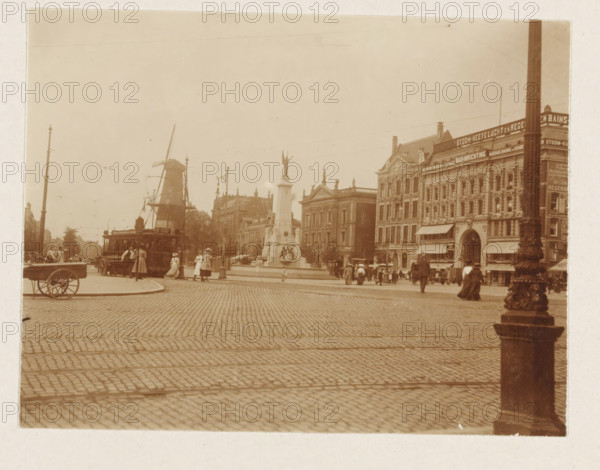 Caland Monument at Van Hogendorpplein in Rotterdam, photograph, Van Hogendorpplein in Rotterdam with Caland Monument, tram line, windmill and bathing facility. Part of the loose-leaf photo album with 39 photos of a trip through the Netherlands, ca. 1907-1910., Rotterdam, c. 1907 - c. 1910, photographer, paper, pasteboard, daylight collodion silver print, height 80 mm x width 110 mm, height 180 mm x width 235 mm, tram, windmill, square, circus, etc. (city(-scape) with figures, staffage), Caland Monument, A wide cobblestone square with a tall ornate column topped by a statue near a central fountain, a large historic building with detailed facades and awnings along one side, a windmill visible in the background, a tram with people standing nearby, several groups of pedestrians in period clothing walking or standing, a wooden cart at the curb, a decorative post or column in the foreground, and streetlamps and overhead wires crossing the scene.