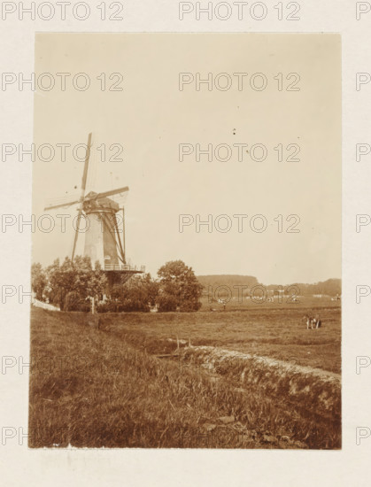 Windmill of the Schieveen polder near Overschie, photograph, Meadow with the upper windmill of the Schieveen polder near Overschie. Part of the loose-leaf photo album with 39 photos of a trip through the Netherlands, ca. 1907-1910., Netherlands, c. 1907 - c. 1910, photographer, paper, pasteboard, daylight collodion silver print, height 110 mm x width 80 mm, height 180 mm x width 235 mm, meadow, pasture, windmill in landscape, Rotterdam, Overschie, Period, 1907 - 1910, A sepia-toned photograph showing a traditional windmill with large sails on the left side, surrounded by trees and a fence at its base, an open grassy field extending from the foreground toward the right, a group of people standing in the field near the middle ground, scattered distant buildings and trees along the horizon, and an expanse of clear sky occupying the upper portion of the image.