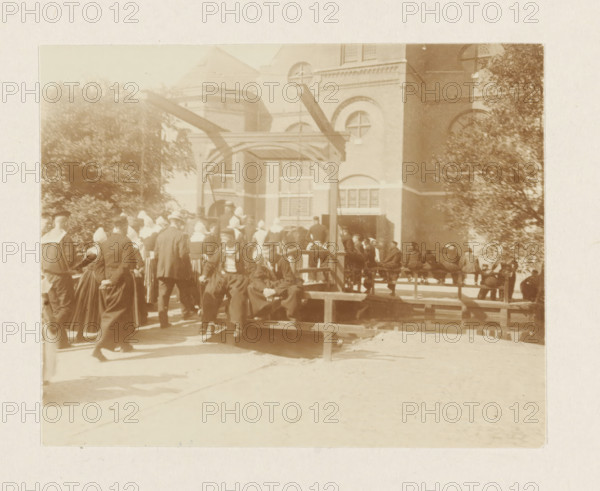 Churchgoers in Volendam, photograph, Men and women in traditional costume cross the drawbridge to the St. Vincentius Church in Volendam. Part of the loose-leaf photo album with 39 photos of a trip through the Netherlands, ca. 1907-1910., Volendam, c. 1907 - c. 1910, photographer, paper, pasteboard, daylight collodion silver print, height 80 mm x width 110 mm, height 180 mm x width 235 mm, folk costume, regional costume, church (exterior), going to church, village (city(-scape) with figures, staffage), St. Vincentius Church (Volendam), A sepia-toned outdoor scene shows a group of people gathered around a wooden platform and a simple wooden frame structure in front of a large brick building with arched windows and decorative masonry. Many figures wear long dark garments and white head coverings, while others wear dark suits and hats. Trees with dense foliage flank the scene on both sides. Some people are seated on the platform while others stand or walk nearby. The ground in front of the...