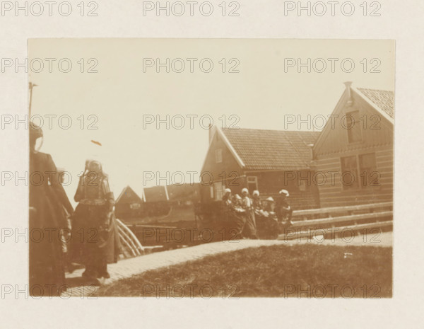 Women and children in Marken, photograph, Children and women in traditional costume on the street near a bridge in Marken. Part of the loose-leaf photo album with 39 photos of a trip through the Netherlands, ca. 1907-1910., Marken, c. 1907 - c. 1910, photographer, paper, pasteboard, daylight collodion silver print, height 80 mm x width 110 mm, height 180 mm x width 235 mm, folk costume, regional costume, village (city(-scape) with figures, staffage), A sepia-toned photograph showing part of a street scene with a tiled-roof house on the right, featuring a gable and visible windows, a low fence or railing in front of the house, and a group of people wearing long garments and head coverings gathered near the fence; on the left edge a person in a dark coat and another figure wearing patterned clothing and a headscarf are partially visible, and a curved paved walkway and grassy area occupy the foreground.