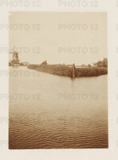 Windmill by the water (former title), Sawmill De Vriendschap in Monnickendam, photograph, View over the water with a windmill and a dolphin. Part of the loose-leaf photo album with 39 photos of a trip through the Netherlands, ca. 1907-1910., Netherlands, c. 1907 - c. 1910, photographer, paper, pasteboard, daylight collodion silver print, height 110 mm x width 80 mm, height 180 mm x width 235 mm, windmill in landscape, Monnickendam, A sepia-toned photograph showing a calm body of water in the foreground with gentle ripples, a narrow grassy peninsula extending into the water toward the middle distance, a wooden pile structure at the tip of the peninsula, reeds and low vegetation along the shoreline, a windmill and a few low buildings visible on the far left shoreline, and an open pale sky occupying the upper portion of the image.