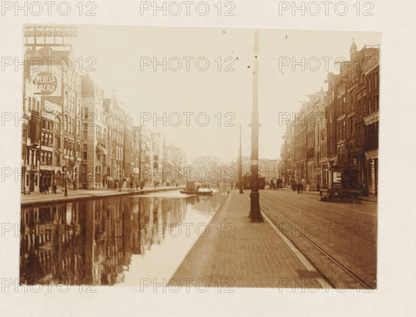 Rokin in Amsterdam, photograph, View over the Rokin in Amsterdam. Part of the loose-leaf photo album with 39 photos of a trip through the Netherlands, ca. 1907-1910., Amsterdam, c. 1907 - c. 1910, photographer, paper, pasteboard, daylight collodion silver print, height 80 mm x width 110 mm, height 180 mm x width 235 mm, canals, waters (in city) (city(-scape) with figures, staffage), street (city(-scape) with figures, Rokin, A sepia-toned street scene shows a canal running alongside rows of multi-story buildings with narrow facades and varied rooflines, their reflections visible in the water; a paved sidewalk and tram tracks run parallel to the canal with a tall streetlamp in the foreground; small boats are moored on the canal and a horse-drawn carriage is visible on the street; people stand and walk along the sidewalks and storefronts line the buildings on both sides.