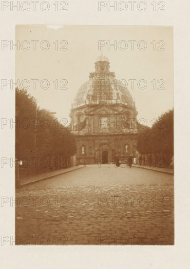 Basilica of Our Lady of Scherpenheuvel, photograph, Basilica of Our Lady of Scherpenheuvel. Part of the loose-leaf photo album with 39 photos of a trip through the Netherlands, ca. 1907-1910., Scherpenheuvel, c. 1907 - c. 1910, photographer, paper, pasteboard, daylight collodion silver print, height 110 mm x width 80 mm, height 180 mm x width 235 mm, church (exterior), A sepia-toned photograph of a wide cobblestone street leading toward a large domed stone building centered in the frame, flanked on both sides by rows of tall, dark trees that create a corridor effect; the dome features a smaller cupola on top and decorative architectural details on its lower portion, while the building's facade shows arched openings and ornamentation; a few small human figures are visible near the building entrance, and the scene has an overall aged, slightly faded appearance.