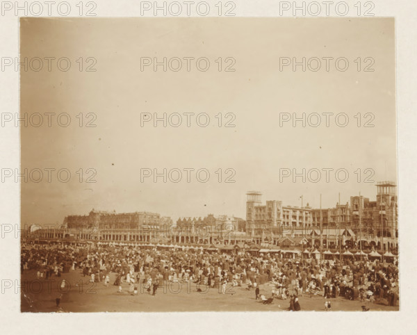 Beach at Scheveningen, photograph, Beach at Scheveningen. Part of the loose-leaf photo album with 39 photos of a trip through the Netherlands, ca. 1907-1910., Scheveningen, c. 1907 - c. 1910, photographer, paper, pasteboard, daylight collodion silver print, height 80 mm x width 110 mm, height 180 mm x width 235 mm, beach (as place of recreation), A sepia-toned photograph showing a crowded beach scene with many people scattered across the sand, many wearing long dresses and hats, some sitting or standing and a few walking; behind the beach there is a long row of multi-story buildings with ornate facades and arched colonnades, including taller structures with rooftop frameworks and towers, and a line of small beach huts or tents in front of the buildings; the sky above is mostly clear and occupies a large portion of the image.