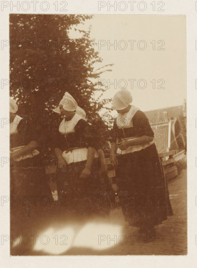 Volendam, photograph, Women in traditional costume in Volendam. Part of the loose-leaf photo album with 39 photos of a trip through the Netherlands, ca. 1907-1910., c. 1907 - c. 1910, photographer, paper, pasteboard, daylight collodion silver print, height 110 mm x width 80 mm, height 180 mm x width 235 mm, folk costume, regional costume, Three women stand side by side wearing dark dresses with light-colored aprons and intricately patterned caps covering their hair and part of their faces, one woman holds a shallow bowl against her midsection while another's hand rests near her waist, behind them a large leafy tree fills the left background and a slatted roof structure is visible on the right, the photo has a sepia tone and shows worn edges and a bright overexposed area near the bottom.