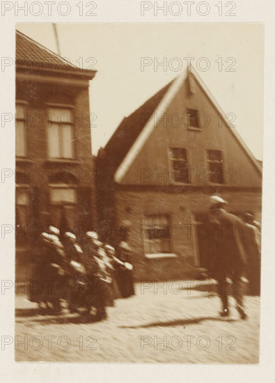 Children in traditional costume, photograph, possibly in Volendam (blurred image). Part of the loose-leaf photo album with 39 photos of a trip through the Netherlands, ca. 1907-1910., Volendam (possibly), c. 1907 - c. 1910, photographer, paper, pasteboard, daylight collodion silver print, height 110 mm x width 80 mm, height 180 mm x width 235 mm, folk costume, regional costume, A sepia-toned photograph showing the façades of two adjacent brick buildings with pitched roofs and multiple windows, one building having a triangular gable. Blurred figures in dark clothing and light-colored caps are moving along the street in the foreground, forming a procession-like group, while a taller, indistinct figure stands to the right. The cobbled street and the shadow cast across it are visible, and the image has soft focus with motion blur and some speckling on the surface.