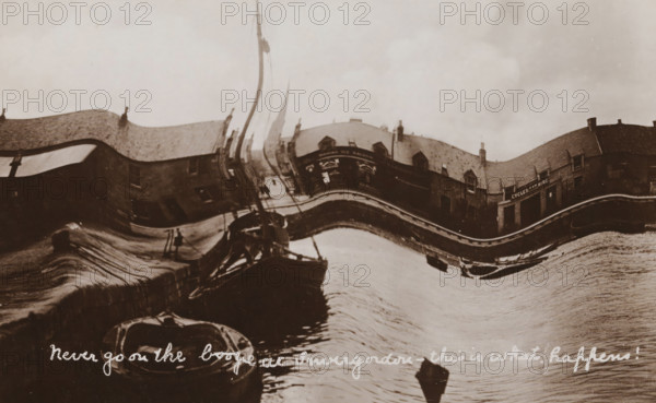 Never go on the boogie at Invergordon - this is what happens!, Davidson's Real Photographic Series, photograph, picture postcard, Invergordon, publisher: Davidson Brothers, London, 1905 - 1920, Davidson Brothers, photographer, publisher, paper, baryta paper, daylight gelatin silver print, height 84 mm x width 136 mm, harbour, drunkenness, A distorted sepia-toned waterfront scene with curved, wavy reflections; a row of buildings with pitched roofs and chimneys along the shoreline, some windows visible and a storefront with a rectangular entrance; a tall mast or pole rising near the center with rigging and fabric suggesting a sail; a narrow quay or stone edge running along the water with small boats moored alongside, one boat bow visible in the foreground and another partially obscured; rippled water surface reflecting the buildings and mast, creating stretched, undulating shapes; an overcast sky above.