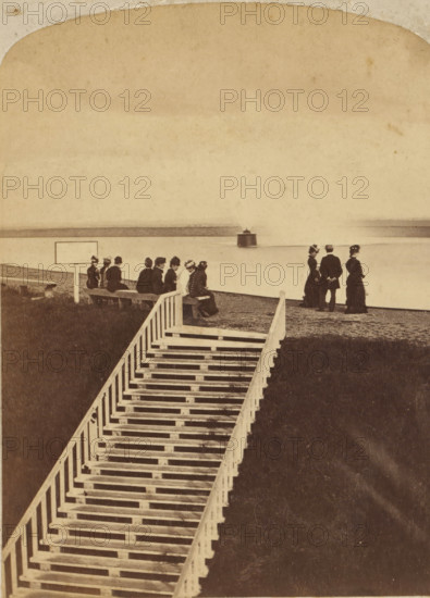 479 mt. Hope Reservoir and Fountain, Rochester New York, stereograph, photographer: George M. Monroe, United States of America, c. 1870 - c. 1880, North America, paper, pasteboard, albumen print, height 120 mm x width 71 mm, height 108 mm x width 176 mm, Rochester (New York), A sepia-toned scene showing a long wooden stairway with white railings leading down through grass toward a shoreline, where a group of people sit on benches near the water and another small group stands facing the water; the seated figures wear dark clothing and hats and are arranged along a low wall, while the standing figures include two women in long dresses and a man in a dark suit and hat; the shoreline is calm and a small rounded structure sits on the water near the horizon, with a faint distant landmass across the water and a wide pale sky above.