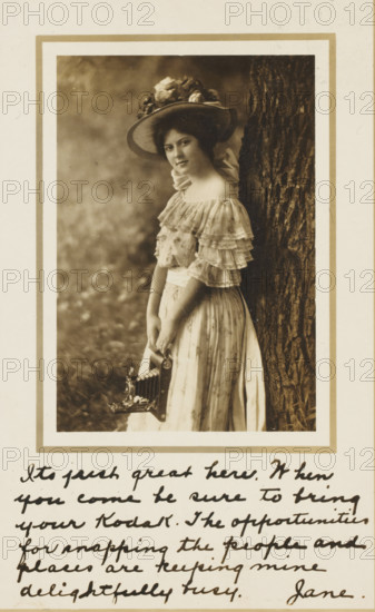 Advertisement for Kodak, picture postcard, stamp: '1909', inscription: 'Its just great here. When you come be sure to bring your Kodak. The opportunities for understanding the people and places are keeping mine delightfully busy. Jane.', United States of America, printer (person): Eastman Kodak, in or before 1909, photographer, printer (person), North America, paper, gelatin silver print, developing gelatin silver print, height 100 mm x width 150 mm, Person, A sepia-toned portrait of a young woman standing beside a textured tree trunk, wearing a wide-brimmed hat adorned with large flowers and a dress with multiple ruffled layers at the bodice and short sleeves, a sash at the waist, and a long skirt; she is facing slightly toward the camera with a neutral expression and holding an old-fashioned camera by its handle near her skirt, all set within a bordered frame on a light background with cursive handwriting visible below the portrait.
