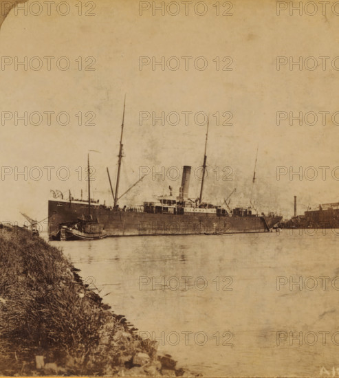 A Mailboat, View of a mailboat at the Handelskade in Amsterdam, Views and landscapes in Europe, Africa and Asia, Geographical Sculpture Gallery, collected by MJ Broekhuijsen, Headmaster of a school in Amsterdam, stereograph, Numbered A12 in the lower right corner of the photo., annotation: 'The 'Burgemeester Den Tex' of the Maatschappij Nederland at the breakwater, at the Handelskade in Amsterdam. In the foreground on the left: a hay barge.', Amsterdam, publisher: Merkelbach & Co., compiler (person in literature): Martien Johan Broekhuijsen, c. 1905, photographer, compiler (person in literature), pasteboard, paper, albumen print, height 87 mm x width 176 mm, Part of a group of 52 stereophotos used in primary education as a supplement to school charts. The entire series consisted of 274 items in 10 categories (A to K)., steamship, motorship, A large vintage steamship sits low in calm water with tall masts and a central funnel rising above a deck crowded with rigging and structures, a sm...