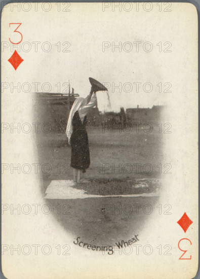Screening Wheat, Three of Diamonds with woman separating wheat from chaff, photograph, playing card, Playing card from a box of 53 playing cards with autotype images of Native Americans. The contents include a complete set of playing cards including a joker., photographer: Adam Clark Vroman, United States of America, publisher: Lazarus & Melzer, Los Angeles (city), c. 1900, North America, paper, autotype, height 88 mm x width 63 mm, A vertically oriented playing card with rounded corners and a worn off-white background, featuring a faded oval vignette in the center that shows a person standing on a platform near water, wearing a dark skirt and a light shawl draped over the head and shoulders, holding a large conical object above their head with material falling from it; distant low buildings and a flat waterfront appear on the horizon within the vignette, and red diamond suit symbols and numerals appear near the corners of the card.