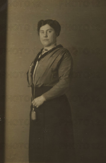 Portrait of Aagje Nieuwmeijer, quick photo, Agatha Jacoba ('Aagje') Nieuwmeijer, profession unknown, wife of Jacobus Johannes Griss, teacher of Dutch Language and Literature at a secondary school in Rotterdam, lived in Rotterdam at the time of the photo., address: 'Kalverstraat 155 E, 178', photographer: American Automatic Fotografie, Amsterdam, 1914, paper, baryta paper, gelatin silver print, developing gelatin silver print, height 86 mm x width 52 mm, Person, Aagje Nieuwmeijer, Kalverstraat, A sepia-toned portrait of a woman standing against a plain backdrop, shown from head to below the waist, with dark hair styled and parted, a composed facial expression, wearing a high-collared blouse and a long-sleeved jacket with a darker trim, a long dark skirt, and holding a small cylindrical object suspended from a cord in one hand at waist level.