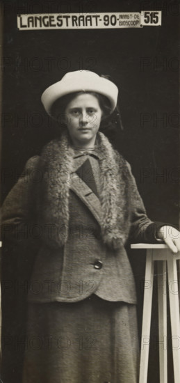 Rapid photographs, snelfoto, photographer: American Tip Top Foto Company, Alkmaar, before 1913-07-08 - after 1917-07, paper, gelatin silver print, developing gelatin silver print, height 83 mm x width 41 mm, A black and white portrait of a woman standing against a dark backdrop, wearing a light-colored hat and a coat with a fur collar, her face visible and expression neutral, one hand resting on a tall wooden stand with vertical slats, the coat showing a button and textured fabric and a skirt extending below, the image framed in a tall narrow format.