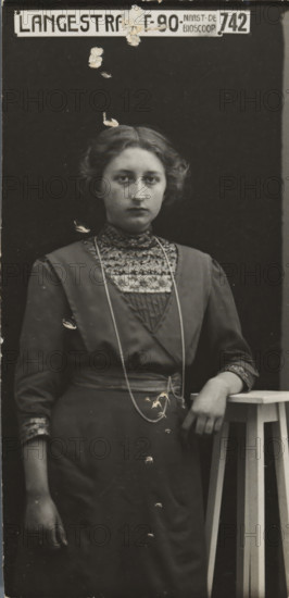 Rapid photographs, snelfoto, photographer: American Tip Top Foto Company, Alkmaar, before 1913-07-08 - after 1917-07, paper, gelatin silver print, developing gelatin silver print, height 77 mm x width 37 mm, A monochrome portrait of a young woman standing against a dark backdrop, shown from head to below the waist, with short wavy hair and a neutral facial expression; she wears a high-collared dress with a patterned yoke and decorative trim at the cuffs, a long thin necklace draped over the front of the dress, and a belt at the waist; her right arm hangs by her side while her left arm rests on a tall wooden stool with visible slatted legs, and there are small areas of surface damage or peeling visible across the photograph, including near the top and over the woman's torso and lower dress; a rectangular sign with block lettering is positioned above her head at the top edge of the image.