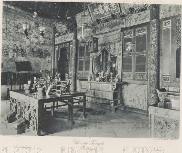 Interior of a Chinese temple, presumably in Kloempang on Sumatra, Kloempang. Chinese Temple, photomechanical print, Part of Album with heliogravures of Sumatra's east coast., number: '12', photographer: Carl J. Kleingrothe, Sumatra, cliché maker: Johann Baptist Obernetter, Munich, in or before 1898, Carl J. Kleingrothe, Johann Baptist Obernetter, photographer, cliché maker, Bavaria, paper, photoaquatint, heliogravure, height 202 mm x width 270 mm, temple, shrine, Hinduism, Buddhism, Jainism, A room filled with ornate wooden furniture and carved panels, showing a large table in the foreground with various vessels and small objects arranged on it, surrounded by wooden benches and chairs. The walls and pillars are covered with intricate painted panels and decorative reliefs, and several hanging glass lanterns and a chandelier descend from the ceiling. Behind the table an elaborately carved cabinet or altar stands against the wall, topped with vases and sculptural ornaments and flanked by ...