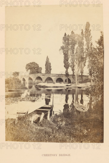 View of the Chertsey Bridge over the Thames, Chertsey Bridge, photograph, page, photographer: attributed to Francis Frith, Surrey, c. 1862 - in or before 1867, Attributed to, paper, albumen print, height 100 mm x width 140 mm, river, Thames, A sepia-toned photograph shows a stone arch bridge spanning a calm river, with multiple rounded arches and a low parapet, tall trees lining the far riverbank rising above the bridge, and reflections of the bridge and trees mirrored in the water; in the foreground there is grassy riverbank with wooden boats moored at the edge, some partially in the water and some alongside simple wooden planks, and water lilies and reeds float near the near shore.