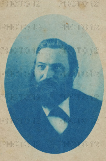 Portrait of man with beard, photograph, Netherlands, 1890 - 1940, photographer, paper, cyanotype, height 128 mm x width 180 mm, historical persons, An oval portrait printed in blue tones on textured beige paper shows a man from the chest upward wearing a dark jacket and a light shirt with a dark tie, his hair combed back and a full beard and mustache, his gaze directed slightly to one side, with subtle paper spots and variations in the blue wash visible across the image.