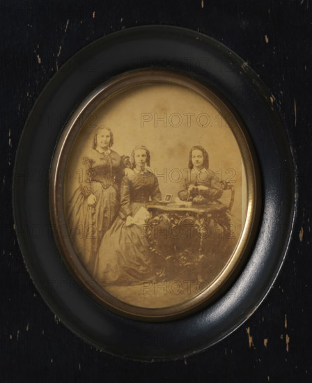 Group portrait of three unknown women around a table, with photos on top, frame, photograph, label: 'Firma Gerard Pisters, (H. Grevers), Steenweg 4, Roermond', frame maker: Firma Gerard Pisters, Roermond (Limburg), c. 1855 - c. 1860, photographer, framemaker, plant material, pasteboard, biological material, inorganic material, imitative material, paper, glass, metal, wood (plant material), imitation leather, albumen print, albumen print in frame, height 74 mm x width 60 mm, height 142 mm x width 120 mm, historical persons not known by name - BB - woman, historical persons depicted in a group, in a group portrait, An oval sepia photograph set in a round dark wooden frame with a slightly worn surface showing small chips and scratches; inside the photograph three young women in period dresses posed together, one seated at a small ornate table with a decorative carved surface, one seated beside her holding a folded handkerchief, and one standing with a hand resting on the seated woman's sh...