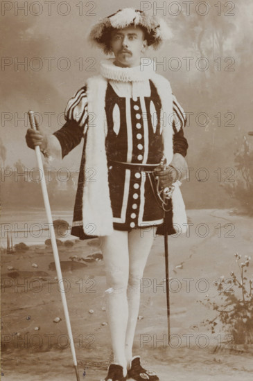 Portrait of Arnold Cornelis Maas Geesteranus in costume, album page, photograph, carte-de-visite, This photo is part of an album., name: 'AC Maas Geesteranus., 23.', inscription: 'Adolphe Zimmermans, de Riemerstraat 21., La Haye., Specialité pour les agrandissements, au charbon inaltérable.', photographer: Adolphe Zimmermans, The Hague, c. 1887, paper, pasteboard, daylight collodion silver print, height 83 mm x width 52 mm, Person, Arnold Cornelis Maas Geesteranus, historical persons, historical costume, A sepia portrait of a person standing full length in a theatrical costume, wearing a wide brimmed hat trimmed with feathers, a ruffled high collar, a dark tunic with bold contrasting trim and round decorative elements down the front, padded striped sleeves, and a long fur-trimmed stole draped over the shoulders; the person holds a long staff in one hand and a sword with an ornate hilt at the waist in the other, and wears light-colored stockings and dark shoes with decorative buckles, s...