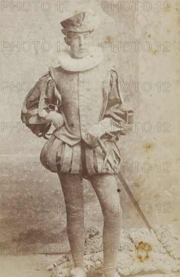 Portrait of a young man in costume, identified as HM Hijmans, album page, photograph, carte-de-visite, This photo is part of an album., inscription: 'J. Goedeljee, Photographer, HRH PRINCE ALEXANDER of the Netherlands, PRINCE of ORANGE, and HRH PRINCESS HENDRIK of the Netherlands, Hoogewoerd 160, near the Plantage.', name: 'HM Hijmans, 53.', photographer: Jan Goedeljee, Leiden, 1860 - 1905, paper, pasteboard, albumen print, height 83 mm x width 52 mm, historical persons, historical costume, A faded monochrome photograph of a person standing and facing slightly to the side, wearing a historical costume with a tall hat, a ruffled collar, a buttoned doublet with padded sleeves, and a skirted garment with vertical panels; one hand rests on the hip while the other holds a long thin sword that extends toward the lower edge of the image; the figure wears tight hose and shoes, and the background shows a mottled textured backdrop and a patterned fabric or rug at the figure’s feet, with visible ...