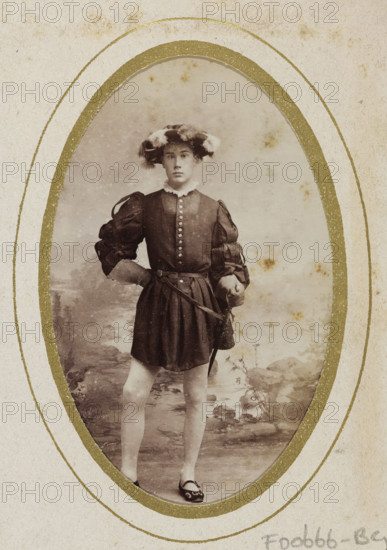 Portrait of a young man in costume with a sword, identified as M. Zijnen de Gier, album page, photograph, carte-de-visite, This photo is part of an album., name: 'M. Zijnen de Gier, 83.', inscription: 'Ant. J. van der Stok, PHOTOGRAPHE, Leiden Rapenburg 13.', photographer: Antonius Joannes van der Stok, Leiden, 1887-01-01 - 1899-12-03, paper, pasteboard, albumen print, albumendruk, height 83 mm x width 52 mm, historical persons, historical costume, hacking and thrusting weapons: rapier, An oval sepia photograph shows a young person standing in a costume with puffed sleeves and a short skirt, hands resting on hips with one hand on a belt holding a sword; the person wears a feathered hat, a high ruffled collar, and flat shoes with decorative details, and the background depicts a painted outdoor scene with rocks and foliage; the photograph is mounted on textured paper with a decorative gold oval border and some age spots visible on both the image and the mount.