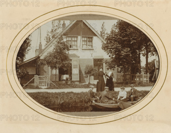 Three men and a woman in a rowboat and two women on shore, with a house in the background, album page, photograph, cabinet photograph, This photo is part of an album., inscription: 'AW van leeuwen, Photographer, Nieuwveen', photographer: AW van Leeuwen, Nieuwveen, 1883 - 1904, paper, pasteboard, daylight collodion silver print, height 137 mm x width 97 mm, rowing-boat, canoe, civic architecture, edifices, dwellings, adult woman (three persons), adult man (three persons), A sepia-toned oval photograph framed by a circular border shows a two-story house with a steep gabled roof, upper windows with curtains, a small porch with an open door, and brick and stucco exterior; trees and shrubs flank the house and a picket gate and fence run along the front; two women in long dresses stand on the lawn near the house while a group of people sit in small rowboats on a canal in front of the lawn, some wearing hats and period clothing.