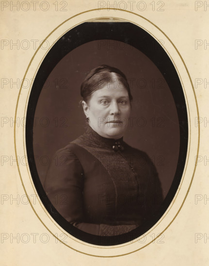 Portrait of a woman, album page, photograph, cabinet photograph, This photo is part of an album., inscription: 'Procedé Inaltérable, Photographie au Charbon, Nestor Schaffers, Photographe, Rue Guinard 9, Gand', photographer: Paul Joseph Nicolas Nestor Schaffers, 1875 - 1896, paper, pasteboard, carbon print, height 134 mm x width 96 mm, historical persons, adult woman, A sepia-toned oval portrait of a woman mounted on a light paper mat with concentric oval borders, showing a woman from the chest up facing forward with a neutral expression, hair parted in the center and pulled back, wearing a dark high-collared dress with a textured front panel and a small decorative clasp at the throat, small earrings, and a plain dark background within the oval frame.