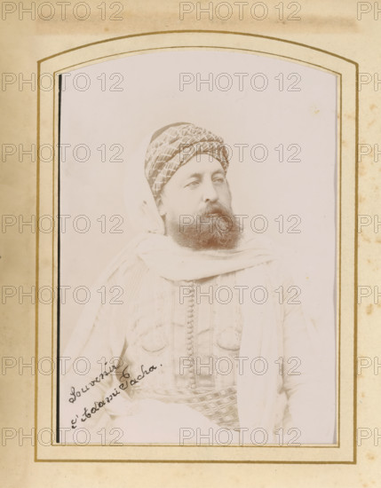 Portrait of a man, album page, photograph, cabinet photograph, This photo is part of an album., inscription: 'Albert, 3 Rue, al Djazira, Tunis, maison fondee, en 1885, Photographe de Son Altesse, le Bey de Tunis, ', inscription: 'A, bert Phot. Tunis', annotation: 'Souvenir, Adami Pacha', photographer: Albert Goupil, 1880 - 1920, paper, pasteboard, daylight collodion silver print, height 134 mm x width 96 mm, historical persons, adult man, A faded portrait of a bearded man wearing a patterned turban and light-colored draped garments set within an arched decorative frame; the man is shown from the torso up, turned slightly to his left with a neutral expression, a textured vest or jacket visible beneath a loose outer cloth, and a patterned sash or belt at his waist; the image and frame both show age with light stains and discoloration around the borders.