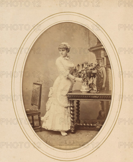 Portrait of a woman standing by a table with flowers, album page, photograph, cabinet photograph, This photo is part of an album., number: '282, 1, 25', inscription: 'Benque & Klary, 33, Rue Boissy d'Anglas, Paris', photographer: Benque et Klary, Paris, 1870 - 1890, Benque et Klary, photographer, paper, pasteboard, albumen print, height 137 mm x height 97 mm, historical persons - BB - woman, adult woman, cut flowers, nosegay, bunch of flowers, A sepia oval portrait of a woman standing beside an ornate wooden table, wearing a light-colored Victorian dress with ruffles, a fitted bodice, and gloves, her hair styled with flowers; she reaches toward a large decorative vase filled with foliage on the table, which also holds a small sculpted cat figure; a carved chair with patterned upholstery sits nearby and a tall carved backdrop or mantelpiece is visible behind the table; the image is framed by concentric oval borders on textured paper.
