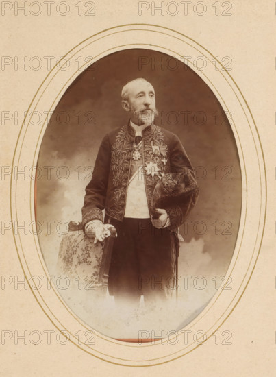 Portrait of Jules Claretie, album page, photograph, cabinet photograph, This photo is part of an album., annotation: '21407, Claretie', inscription: 'R. Dechavannes, Paris', inscription: 'Professeur Stebbing, Photographe hors concours, Boulevard des Italiens et 30, Rue de Grammont, collector's mark: 'Albert Hautecoeus, 12, Boulevard des cap..., photographer: Édouard Stebbing, Paris, 1870 - 1900, Édouard Stebbing, photographer, paper, pasteboard, albumen print, height 137 mm x width 97 mm, Person, Jules Claretie, historical persons, portrait of a writer, An oval-framed sepia photograph shows an older man standing and facing slightly to the side, wearing a dark embroidered jacket with ornate leaflike patterns and a light-colored waistcoat underneath; he has a trimmed beard and mustache and a high collar at his neck. Medals and decorative insignia are pinned to his chest. He holds a fur-trimmed hat in one hand and rests the other hand on the back of a patterned upholstered chair with a fl...