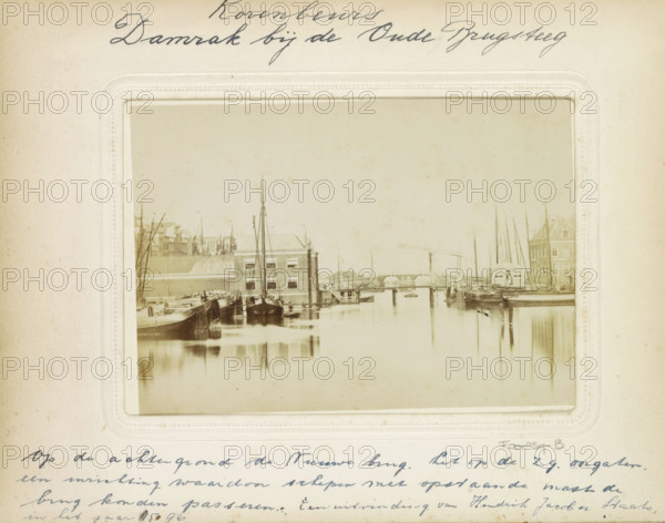 The Damrak. Amsterdam. View of the canal called Damrak., Damrak seen towards the Oudebrug and the Nieuwebrug, with the old Korenbeurs on the left, photograph, cabinet photograph, Part of Photo album with photographs of streets, squares and canals in Amsterdam., photographer: attributed to Andries Jager, Amsterdam, c. 1860 - c. 1875, Attributed to, pasteboard, paper, albumen print, height 107 mm x width 165 mm, prospect of city, town panorama, silhouette of city, canals, waters (in city), Exchange, Bourse, Damrak, Korenbeurs (Amsterdam), Oudebrugsteeg, A sepia-toned photograph mounted on a cream-colored page shows a calm canal scene with rowed and moored boats along both sides, tall thin masts rising above the vessels, a rectangular brick building with multiple windows at the water's edge, a low bridge spanning the canal, reflections of boats and structures in the still water, and surrounding quay structures and a larger building with a pitched roof visible near the right side.