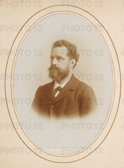 Portrait of a man with mustache and beard, album page, photograph, cabinet photograph, This photo is part of an album., inscription: 'HW Wollrabe, Hoffotograaf, The Hague, NOORDEINDE 96.', photographer: Heinrich Wilhelm Wollrabe, Dordrecht, 1880 - 1910, paper, pasteboard, daylight collodion silver print, height 137 mm x width 98 mm, historical persons, beard, mustache, A sepia-toned oval portrait of a man with wavy hair and a full beard, shown from the chest upward and facing slightly to the viewer's left, wearing a dark suit jacket with a visible lapel and a light-colored shirt with a tied neckpiece, set against a plain light background and framed by a paper mat with multiple concentric oval lines and slight edge wear.