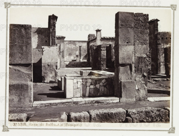 Casa di Sallustio (Pompei), Remains of the House of Sallust in Pompeii, album page, photograph, Part of Photo album with photographs of excavations in Pompeii by Giorgio Sommer., number: '5318', photographer: Giorgio Sommer, Pompeii, c. 1860 - c. 1900, paper, albumen print, height 102 mm x width 139 mm, villa, excavation, archaeology, A black and white photograph of a ruined stone structure showing several weathered walls and columns of varying textures and sizes, with rough blockwork and smoother plastered surfaces; in the foreground there is a low rectangular stone feature with a mosaic-like facing, and uneven paving and large stones lie along the bottom edge; deeper into the scene are fluted column fragments and a narrow open courtyard area framed by tall masonry blocks, shadowed recesses, and broken edges, all captured within a bordered photographic mount.