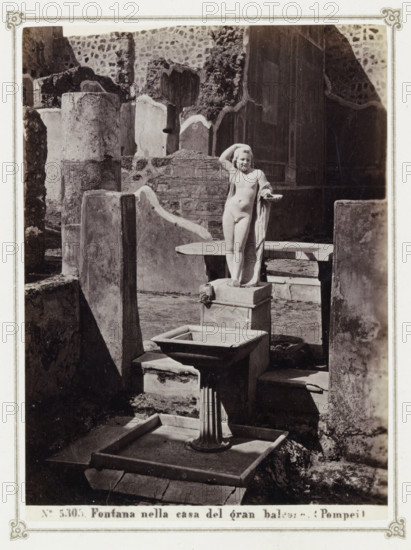 Remains of a house in Pompeii with a statue and a fountain, Fontana nella casa del gran balcone (Pompeii), album page, photograph, Part of Photo album with photographs of excavations in Pompeii by Giorgio Sommer., number: '5305', photographer: Giorgio Sommer, Pompeii, c. 1860 - c. 1900, paper, albumen print, height 140 mm x width 102 mm, piece of sculpture, reproduction of a piece of sculpture, ornamental fountain, excavation, archaeology, A stone courtyard with weathered walls and broken masonry surrounds a central sculpted fountain featuring a small nude child figure standing on a square pedestal with one hand raised to the head and a draped cloth over one arm, a shallow basin mounted on a fluted column stands in front of the pedestal, and scattered stone slabs and low platforms occupy the foreground and background.