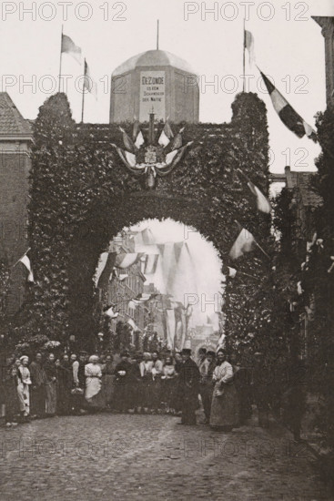Willemstraat., Triumphal arch in the Willemstraat during the April festivities in 1887 in Amsterdam, photograph, cabinet photograph, Part of Photo album with images of the April festivities in 1887 in Amsterdam., Amsterdam, 1887-04, photographer, pasteboard, ink, materials by function, paper, printing ink, collotype, photogravure, height 147 mm x width 100 mm, prospect of city, town panorama, silhouette of city, gate, entrance, April festivities 1887, Willemstraat, A tall archway covered in dense foliage spans a cobblestone street, with a decorative crest and draped fabric at its center and flags on poles rising above; beyond the arch a narrower street is lined with buildings and more hanging banners, and a crowd of people stands beneath and in front of the arch, with some figures in long garments and others in darker coats, their faces and postures visible but not detailed.