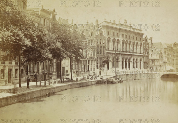 The Voorburgwal with the Post Office. Amsterdam. Le Bureau de Poste., Voorburgwal with the Royal Post Office to the right of the center, photograph, cabinet photograph, Part of a Leporello album with twelve photographs of Amsterdam., photographer: attributed to Andries Jager, Amsterdam, c. 1860 - c. 1880, Attributed to, paper, pasteboard, albumen print, height 94 mm x width 135 mm, prospect of city, town panorama, silhouette of city, canals, waters (in city), post-office, Nieuwezijds Voorburgwal, Post Office (Amsterdam), Royal Post Office, A riverside scene shows a row of multi-story buildings with varied architectural facades and tall narrow windows lining a curved embankment, with a large classical building featuring arched openings and decorative columns standing prominently among smaller neighboring structures; a tree-lined sidewalk runs between the buildings and the water, with leafy trees partially obscuring some facades and streetlamps along the path; a small boat is moored at t...